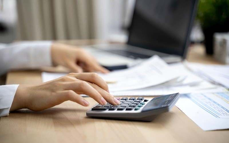 Close-up of a hand using a calculator with papers and a laptop in the background